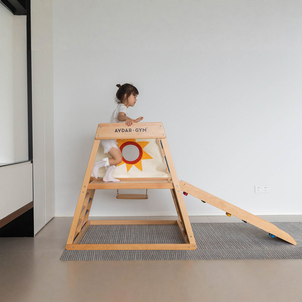 A child playing on a wooden climbing frame which includes a rainbow rack, small climbing rack with cut-outs, climbing rope net, and protective pad.