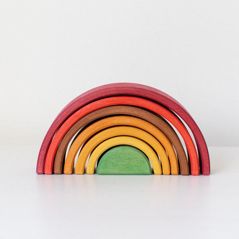 A handcrafted wooden rainbow stacker toy with autumn hues, including pieces in red, orange, yellow, green, and other colors, arranged in a semi-circle on a light background.