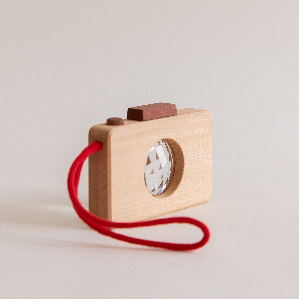 A wooden toy camera with a kaleidoscope lens, featuring a solid wood construction and a red strap.