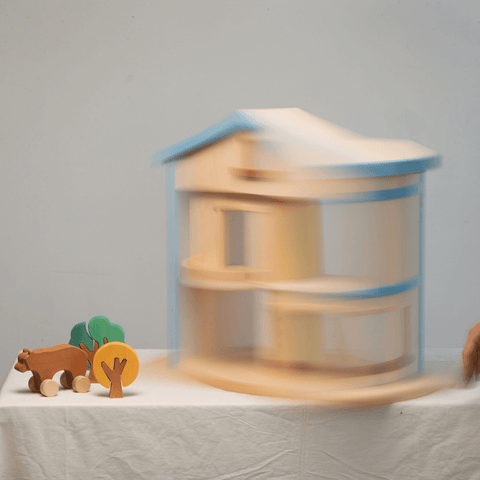 A wooden dollhouse with a blue roof, featuring two stories, an attic, and a balcony on a white table