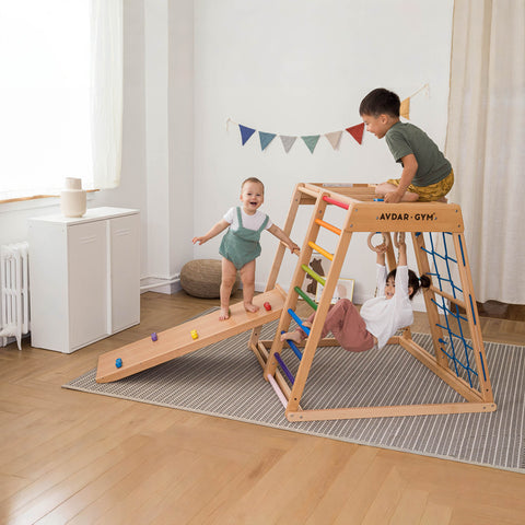 Children playing on a colorful wooden climbing gym set in a bright room, featuring a slide, swing, and ladder.