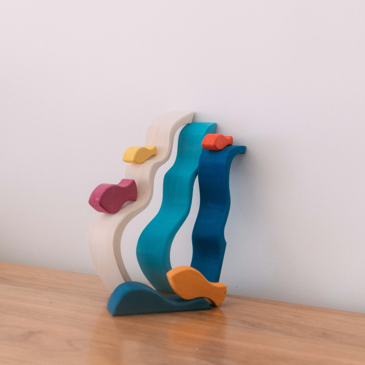 A set of colorful wooden waves with different size and color fishes placed on top, resembling a stackable toy setup on a wooden surface.