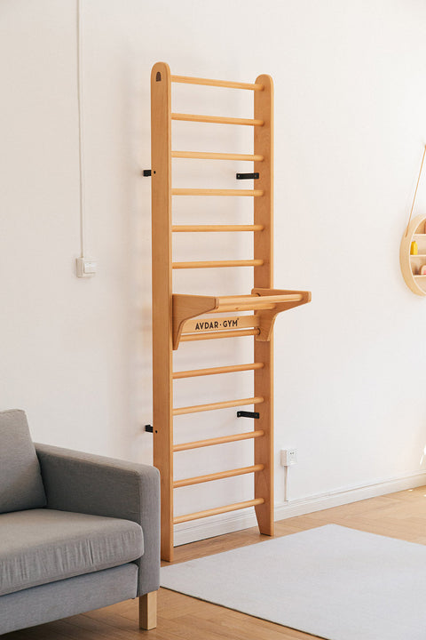 A wooden wall bar set with multiple rungs, mounted on a white wall, with a gray sofa to the left in a home environment.