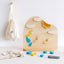 A wooden weather board toy with metal board and magnetic weather pieces, including clouds, sun, and rain symbols, displayed on a white background with a few other toys and a fabric bag in the background.